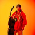 Photograph of Cris Derksen standing, holding a cello, against an orange background.