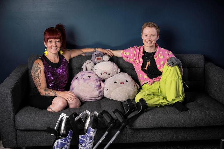 A family portrait of Maxime and Erin, two white Disabled nonbinary neuro non-conforming (neurodivergent) humans, sitting on a couch in bright and vibrant outfits. Between them is a pile of round plushies of various sizes which they affectionately call their kids. Though seemingly inanimate, each of the kids has a unique personality and they are very much alive to Erin and Maxime. Everyone in the photo smiles. Leaning against the couch are Erin’s prosthetic legs and Maxime’s forearm crutches. Photo by Benjamin Laird