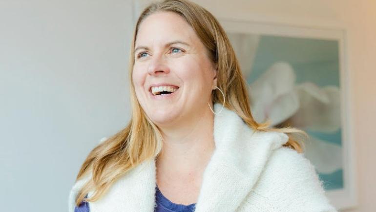 Tara McGee looks out the window of a bright room with a big smile on her face. A soft sweater is draped around her shoulders.