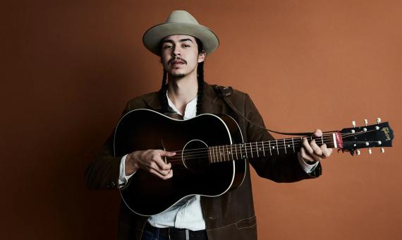 JD Crosstown with a guitar in front of a brown backdrop with soft lighting.