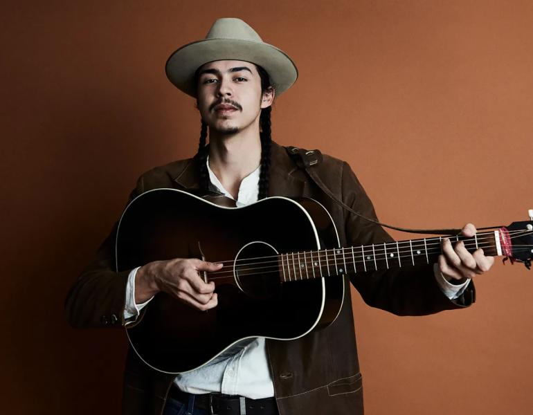JD Crosstown with a guitar in front of a brown backdrop with soft lighting.