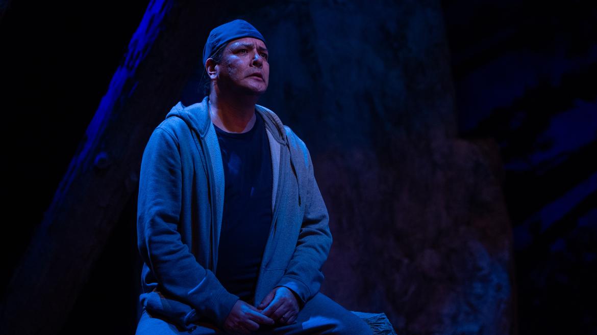 A shot of Jeremy on stage in In The Shadow of the Pines; dark blue light, an outside setting, he looks a little scared